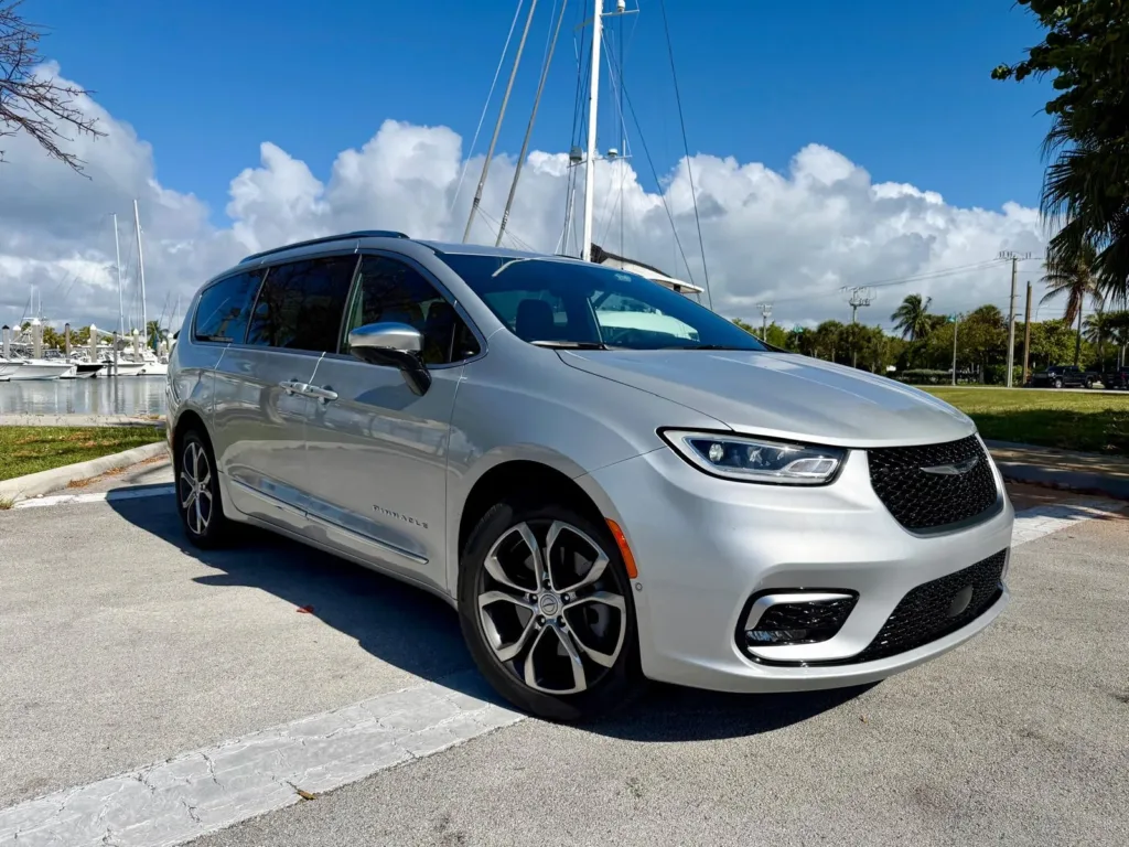 Chrysler Pacifica Pinnacle 2026 plateada junto a una marina con palmeras, vista de tres cuartos delantera