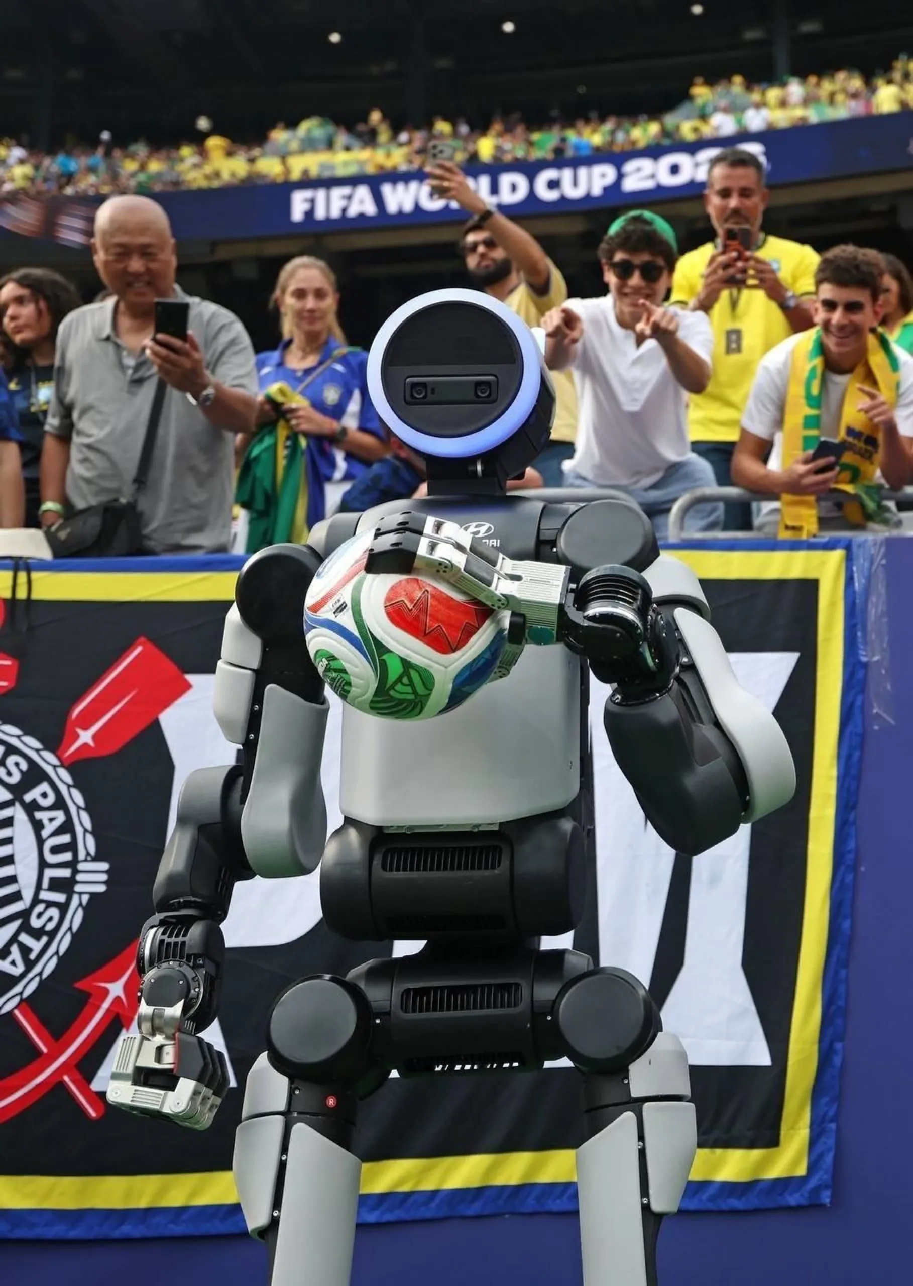 Robot Atlas de espaldas con el balón bajo el brazo frente a hinchas de Brasil que lo graban con sus celulares