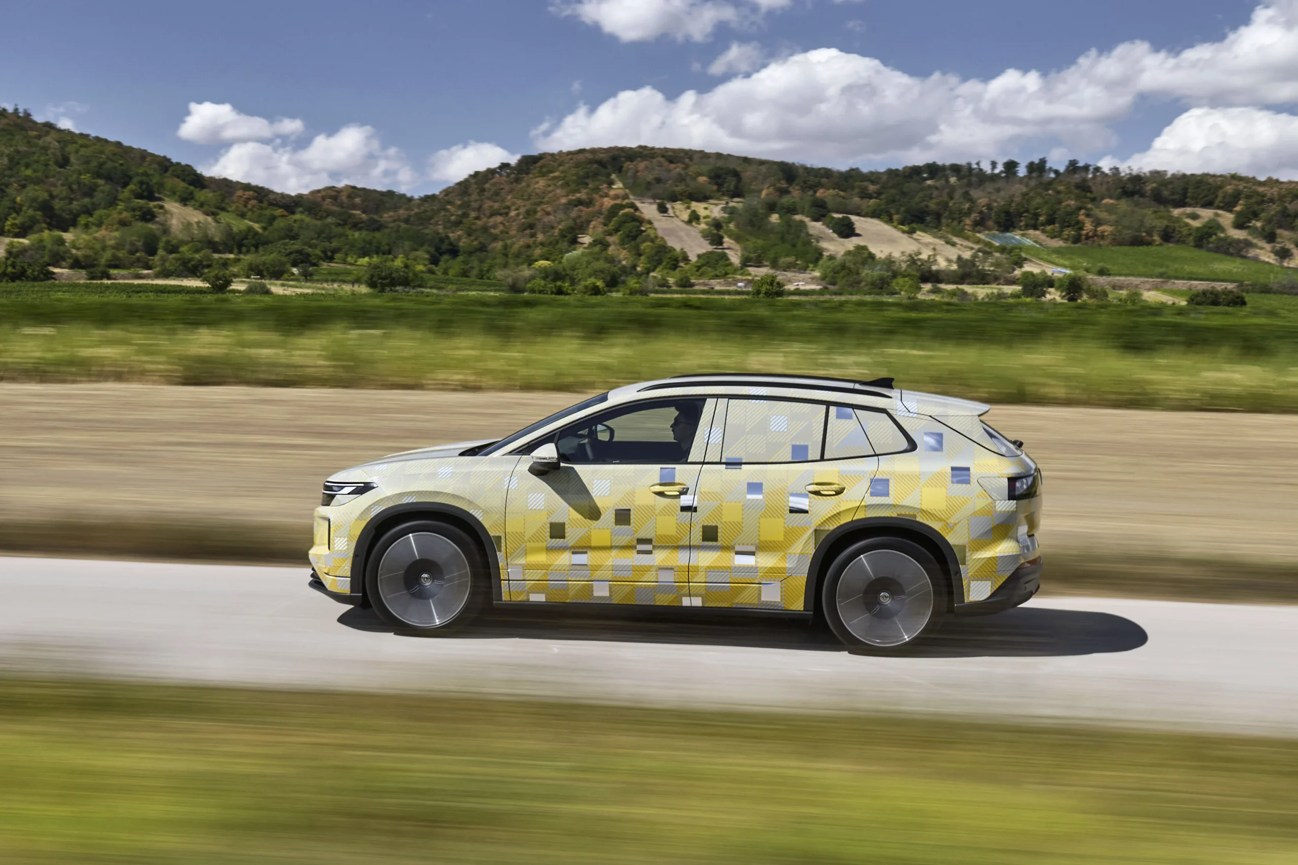 Perfil lateral del prototipo del ID. Tiguan con camuflaje amarillo, en movimiento frente a unas colinas