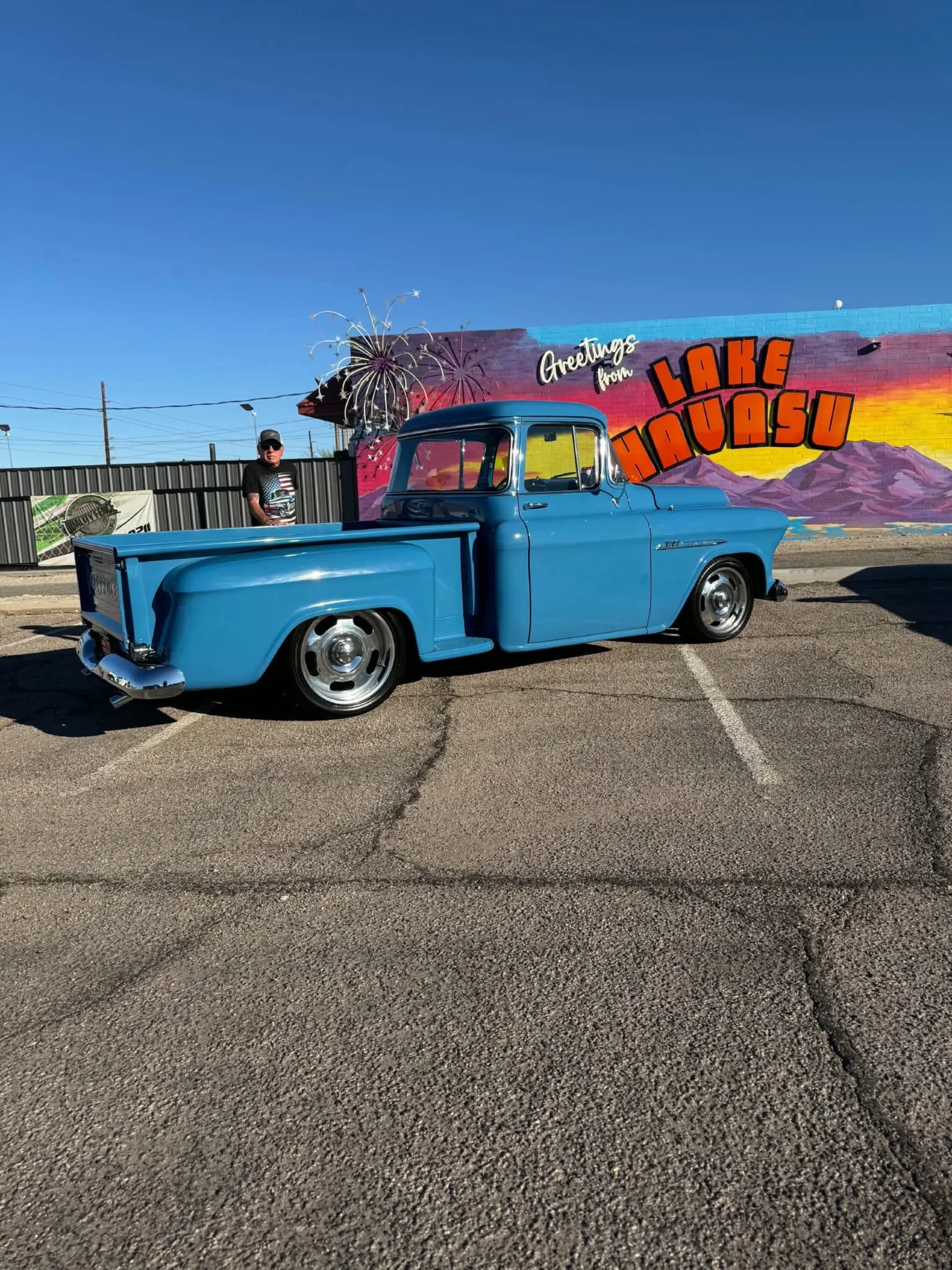 Pickup Chevrolet clásica celeste estacionada frente a un mural de Lake Havasu, con un hombre a su lado