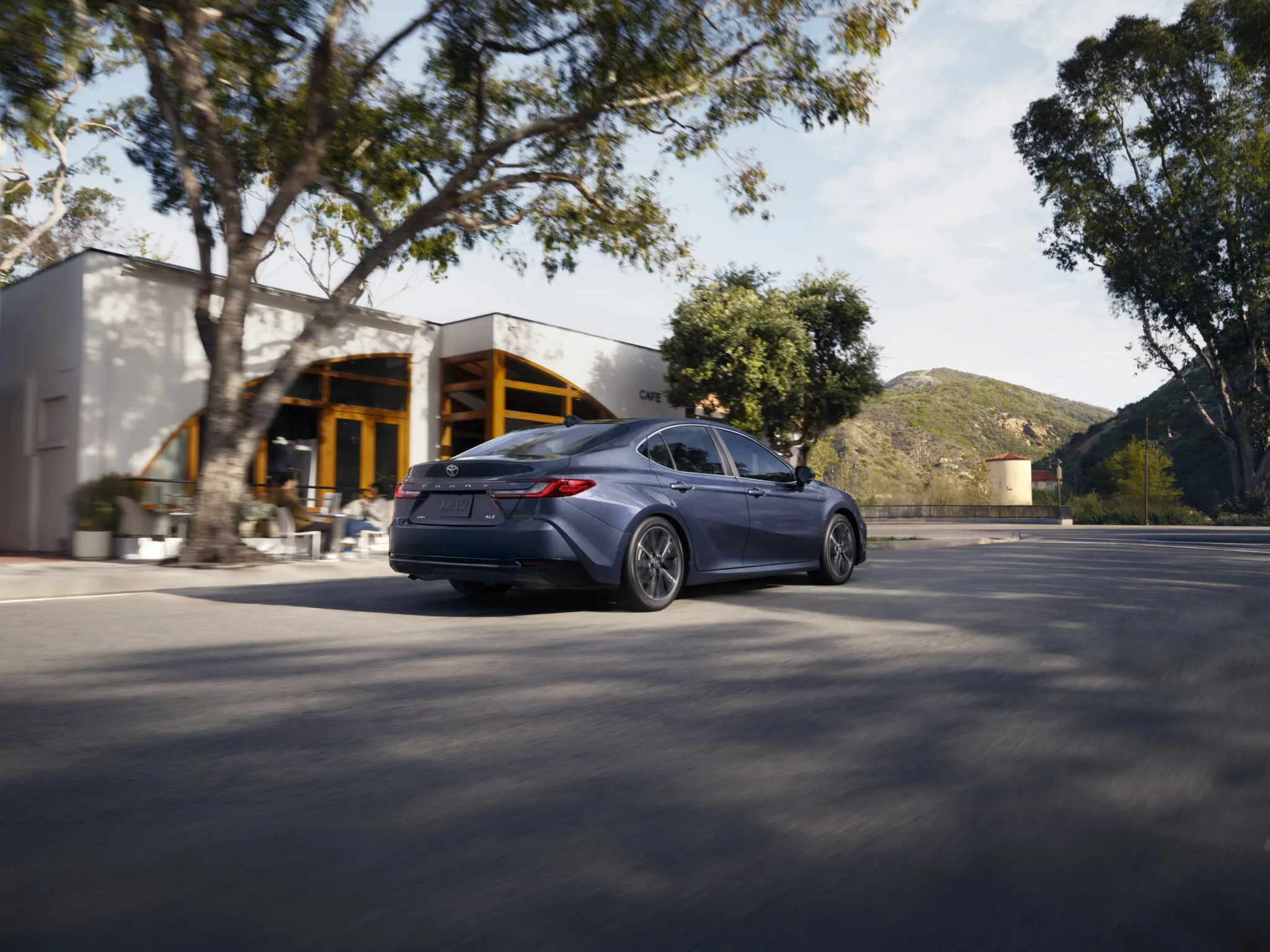 Toyota Camry 2027 gris azulado visto de atrás frente a una cafetería, con una colina al fondo