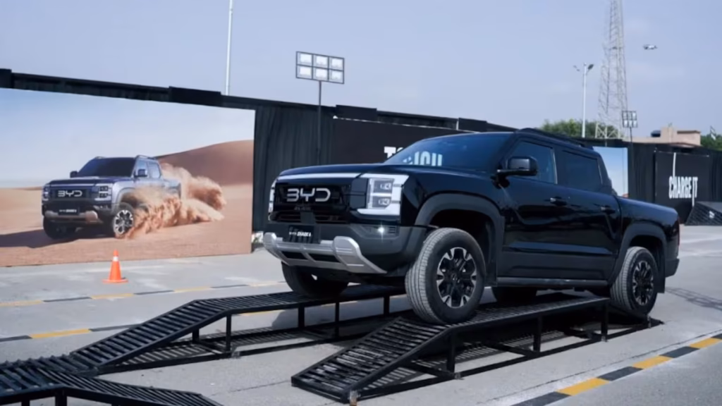 Pickup BYD Shark negra sobre una rampa de exhibición, con una imagen de la camioneta en el desierto al fondo