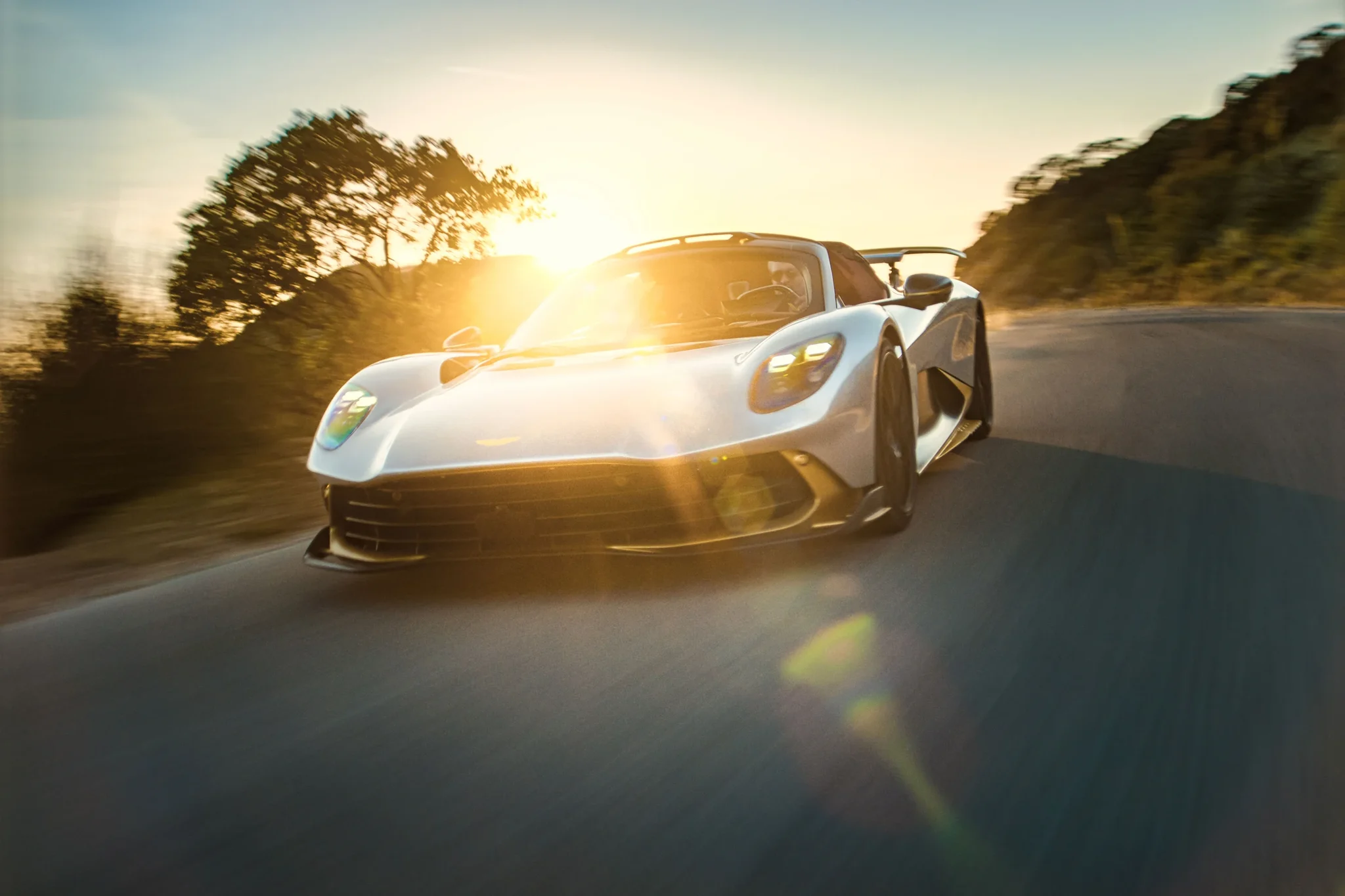 Aston Martin Valhalla Spider de frente al atardecer, con el sol detrás, en una carretera de montaña