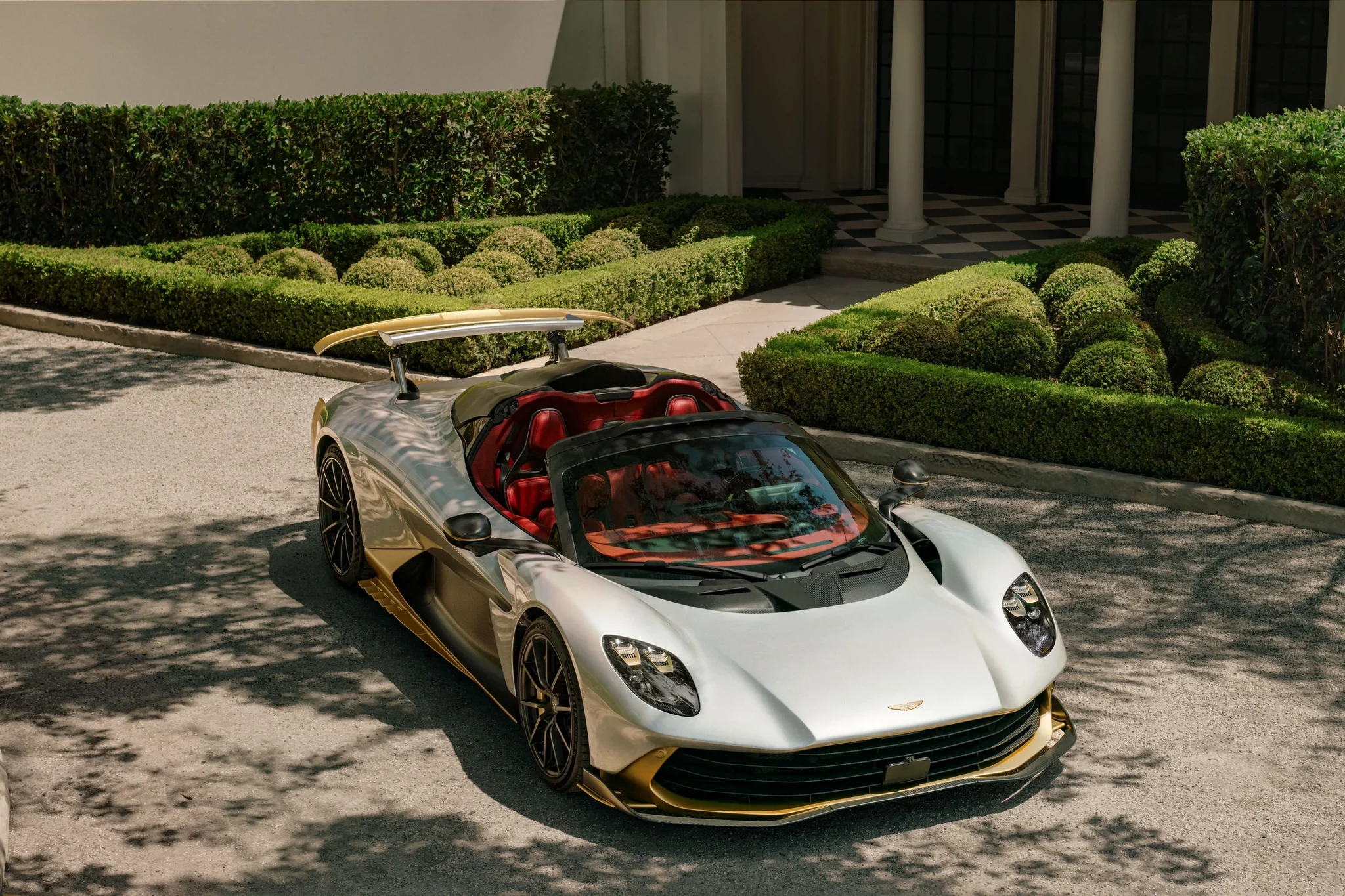 Aston Martin Valhalla Spider plateado con detalles dorados, de tres cuartos frontales y sin techo, frente a un jardín