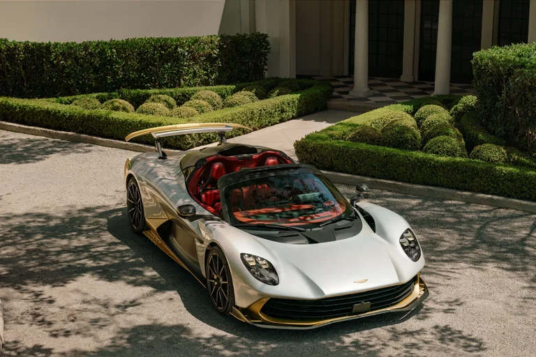 Aston Martin Valhalla Spider plateado con detalles dorados, de tres cuartos frontales y sin techo, frente a un jardín