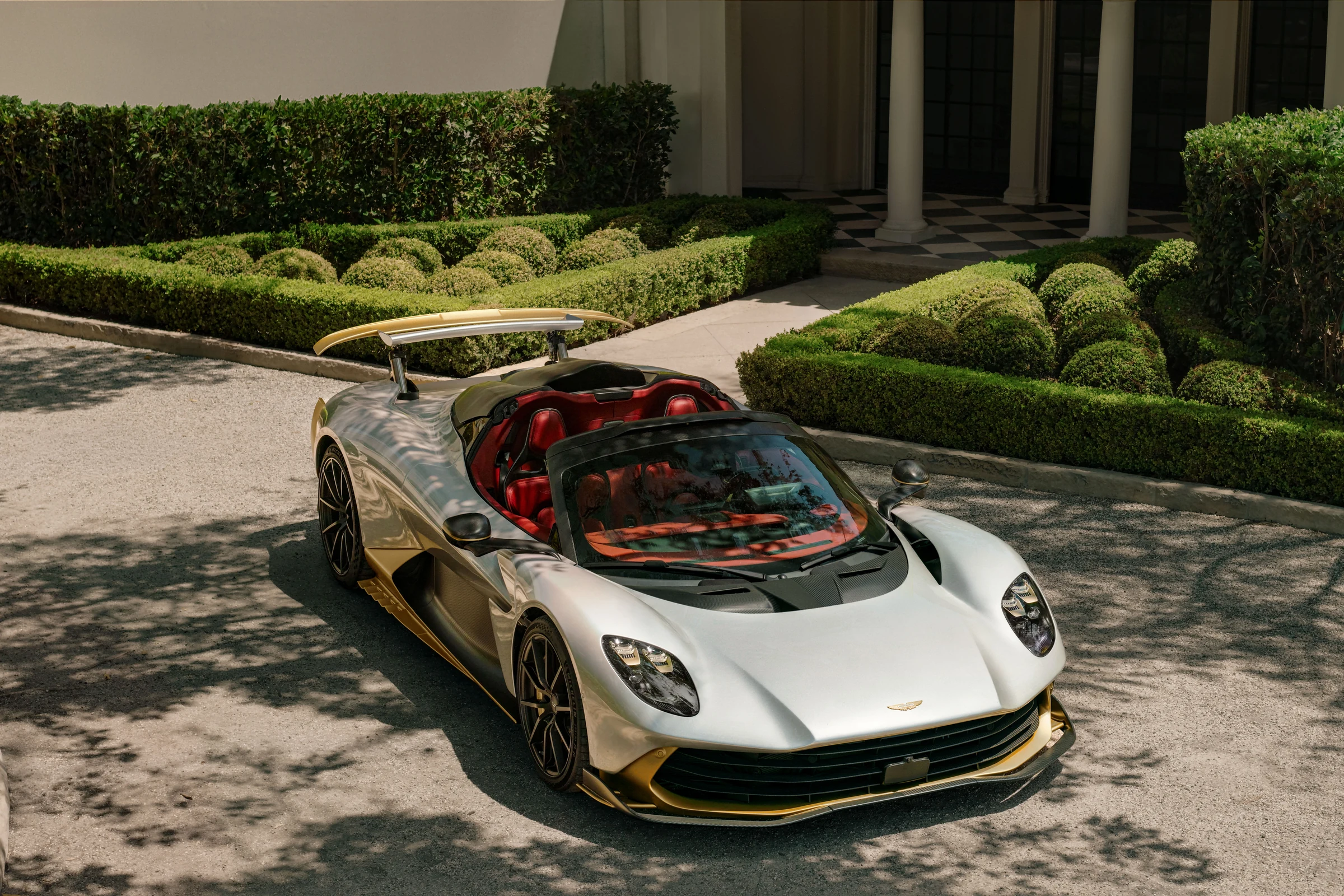 Aston Martin Valhalla Spider plateado con detalles dorados, de tres cuartos frontales y sin techo, frente a un jardín