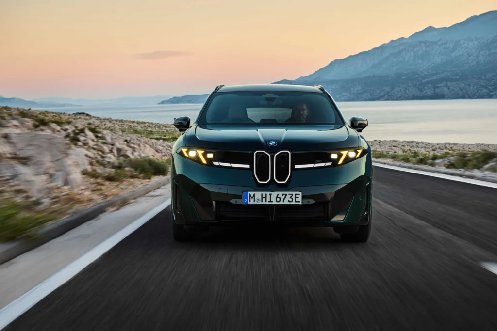 Frente del BMW iX3 M60 xDrive al atardecer en una carretera junto al mar