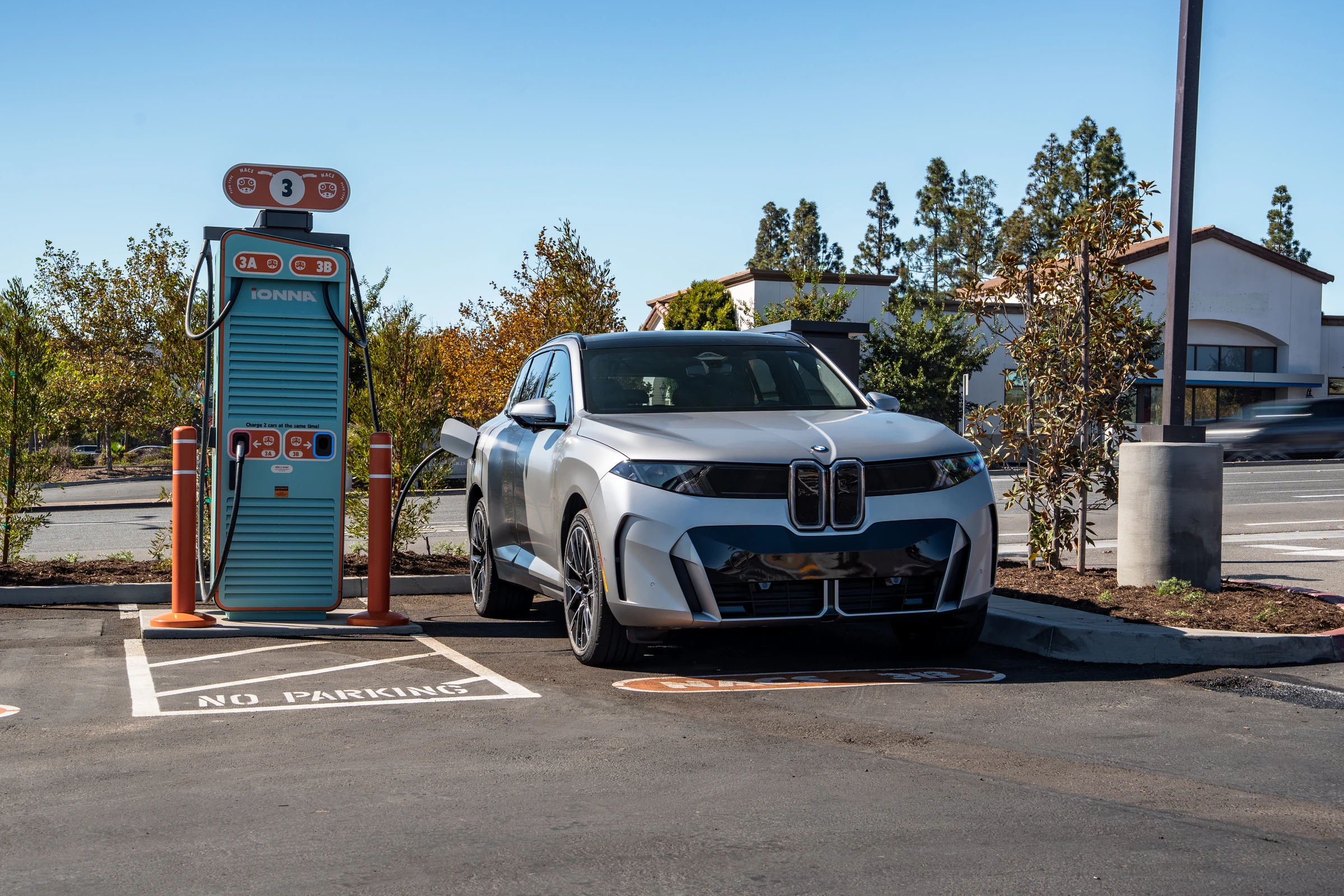 BMW iX5 60 xDrive gris en tres cuartos, estacionado junto a un cargador de IONNA con árboles de fondo