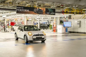 MINI Cooper blanco de frente en la planta de Oxford, bajo un cartel de la marca, con las luces encendidas