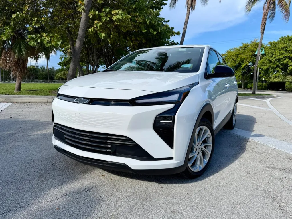 Chevrolet Bolt 2027 blanco, vista frontal tres cuartos, estacionado junto a unas palmeras