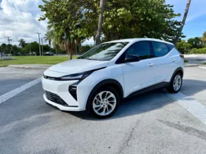 Chevrolet Bolt 2027 blanco visto de frente y de lado, con la calle de fondo