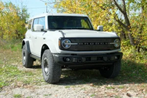Ford Bronco 2027 de cuatro puertas, blanco con techo duro, visto de frente en un camino de grava entre árboles