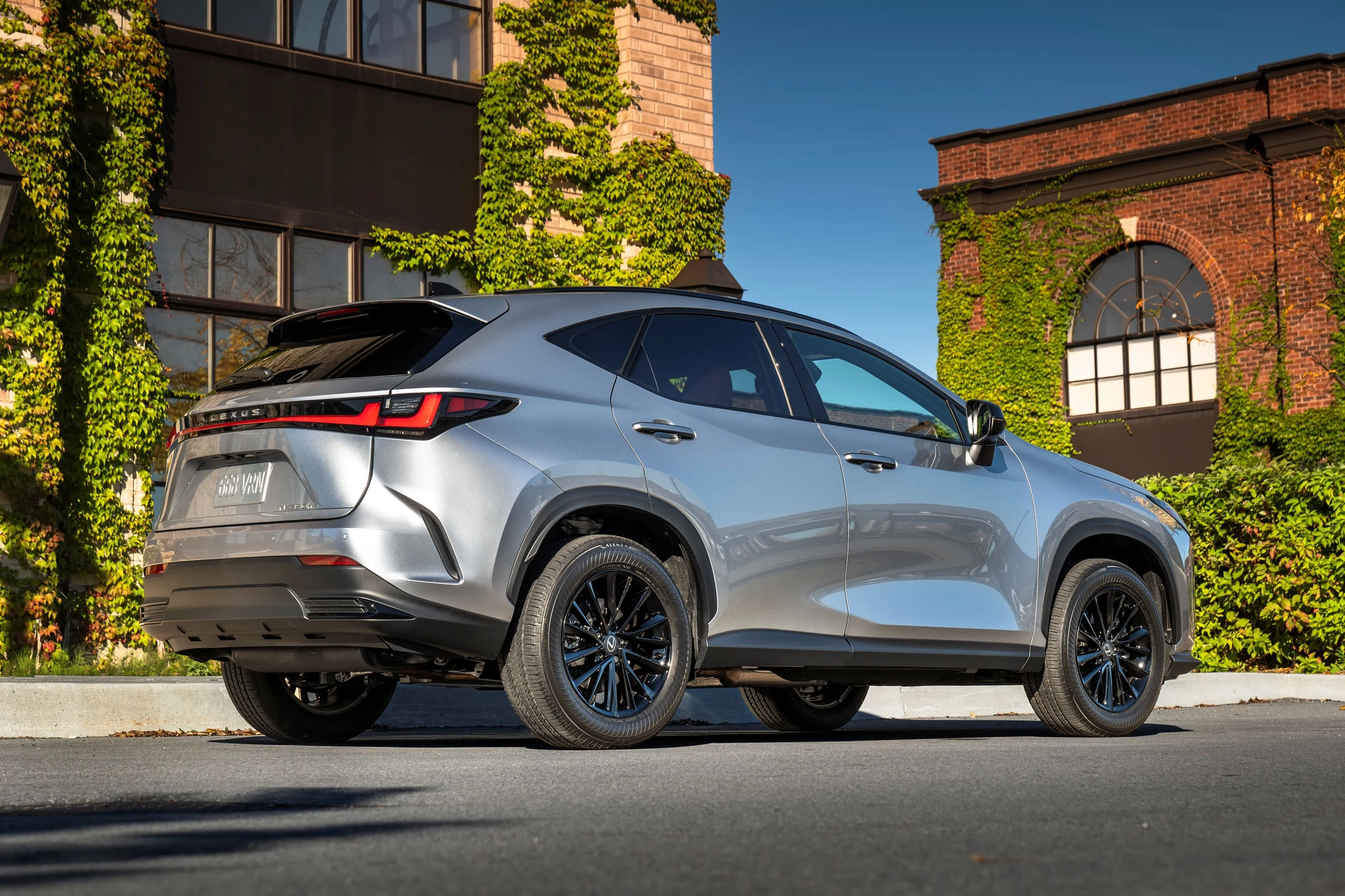 Lexus NX 2027 350h Luxury en color Iridium visto desde atrás, con ruedas negras, junto a un edificio cubierto de hiedra