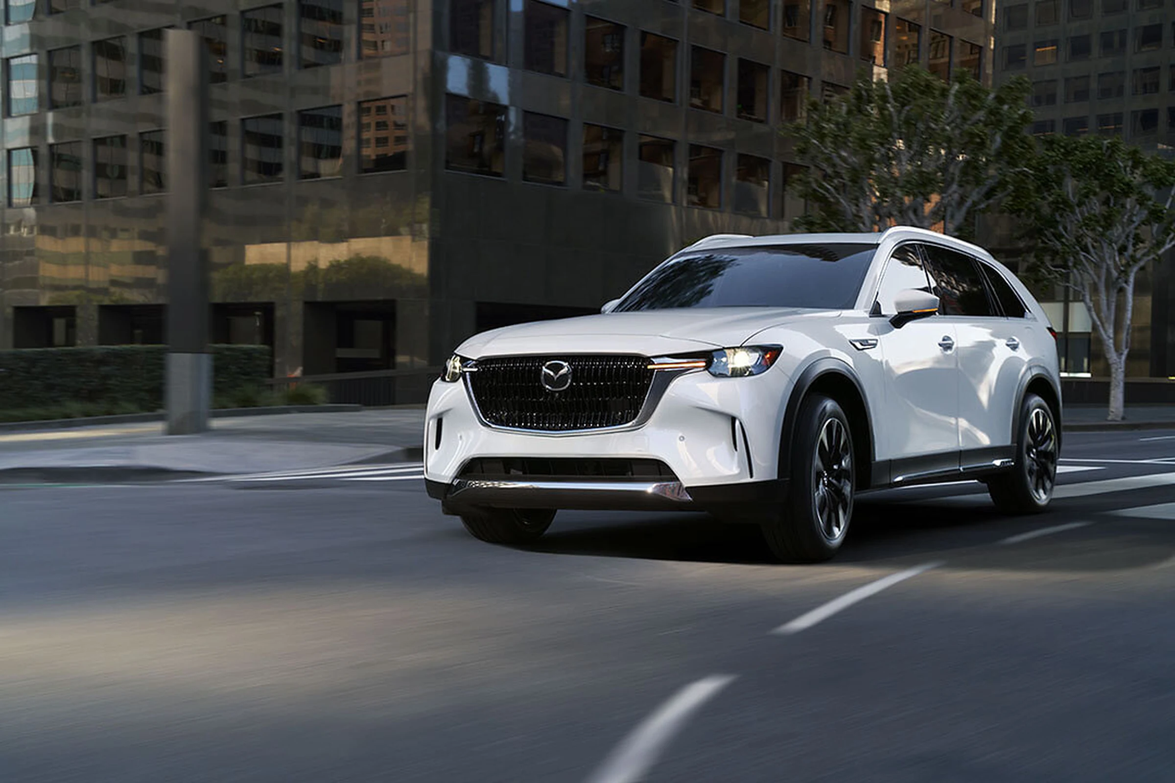 Mazda CX-90 2027 blanco de tres cuartos frontales, en movimiento por una calle entre edificios oscuros