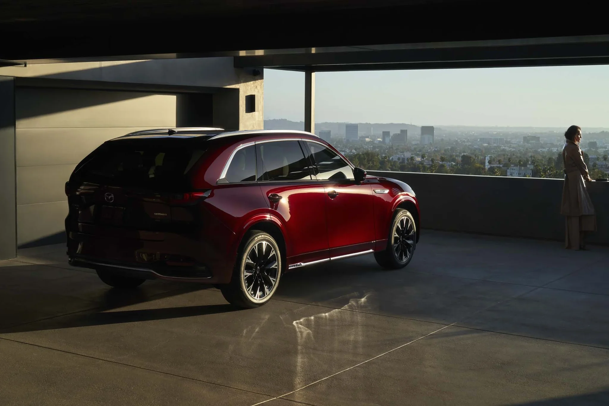 Mazda CX-90 rojo de tres cuartos traseros en un estacionamiento techado, con una mujer mirando la ciudad