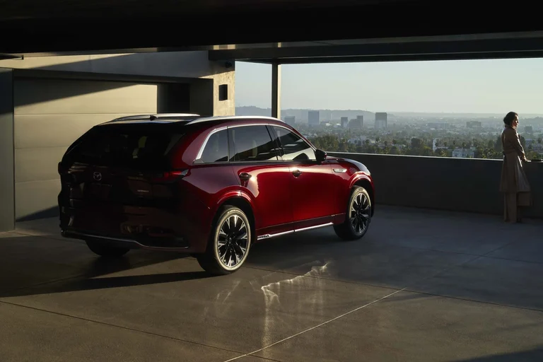 Mazda CX-90 rojo de tres cuartos traseros en un estacionamiento techado, con una mujer mirando la ciudad