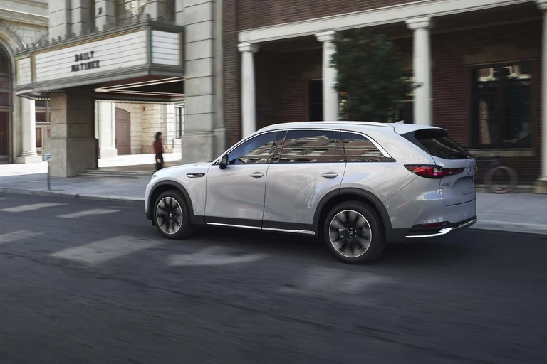 Mazda CX-90 blanco visto de costado y en movimiento frente a un cine de fachada de ladrillo