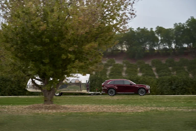 Mazda CX-90 rojo remolcando un tráiler plateado por un camino junto a un parque