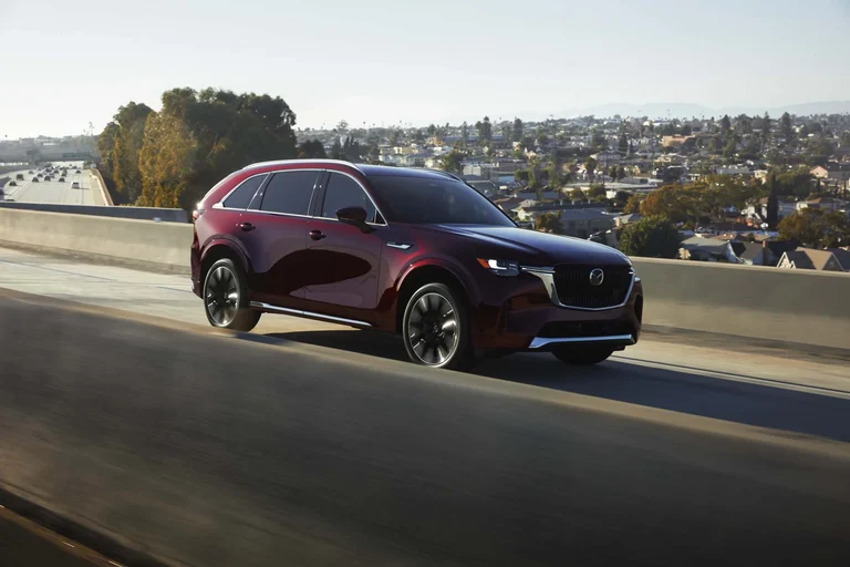Mazda CX-90 rojo de tres cuartos frontales recorriendo un paso elevado con la ciudad al fondo