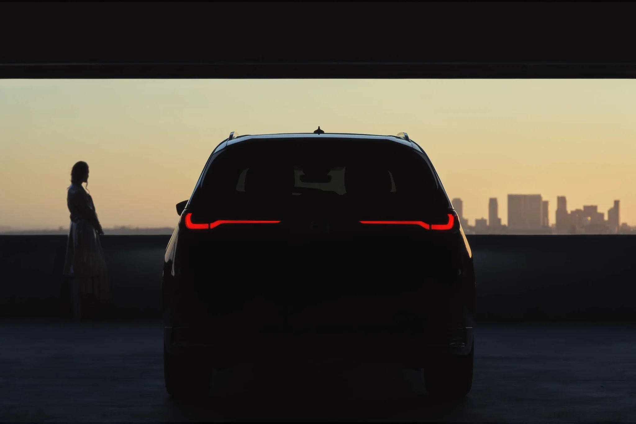 Parte trasera del Mazda CX-90 en penumbra, con una mujer y el horizonte de la ciudad al atardecer