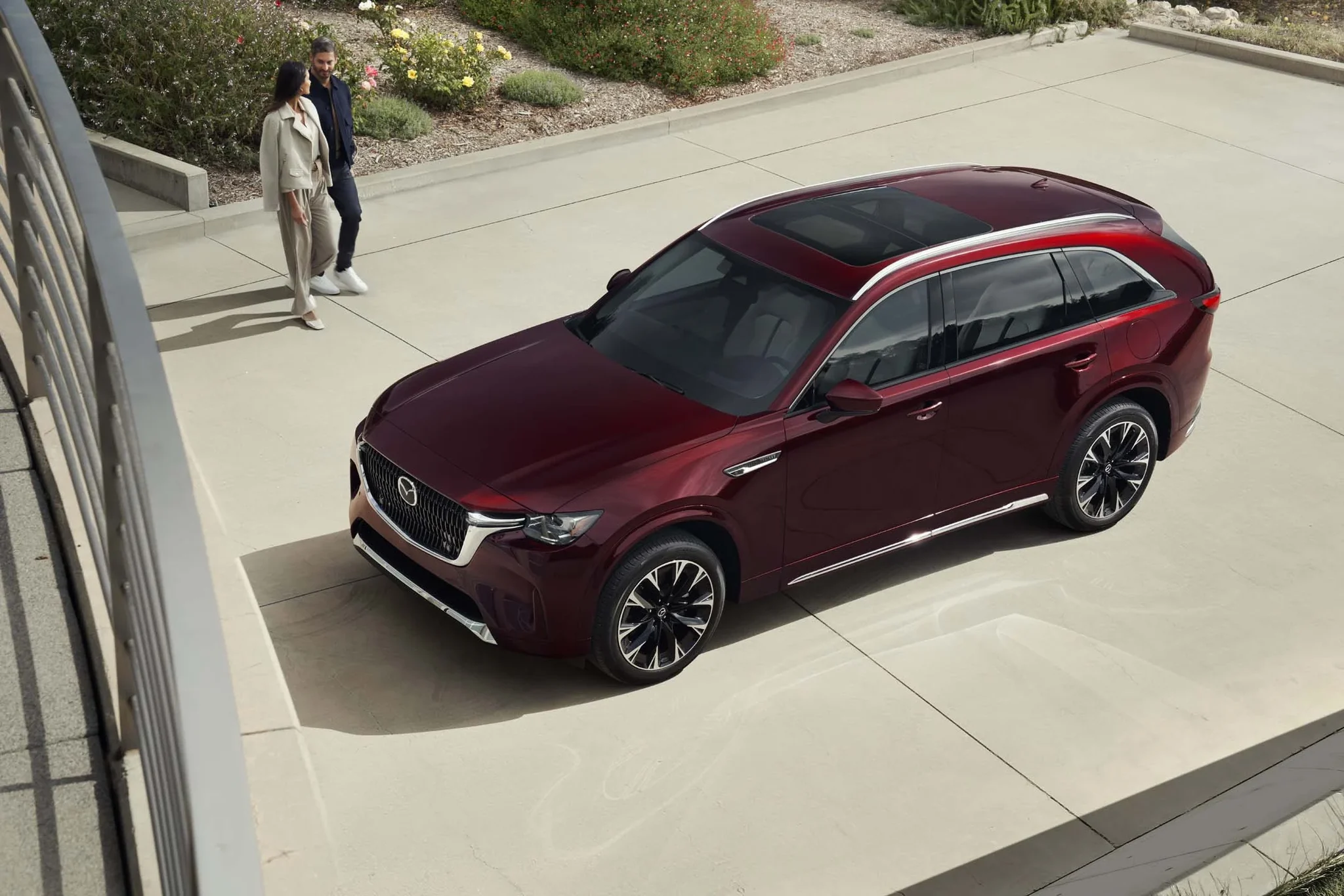 Vista aérea de un Mazda CX-90 rojo detenido en una plaza de concreto, con una pareja caminando al fondo