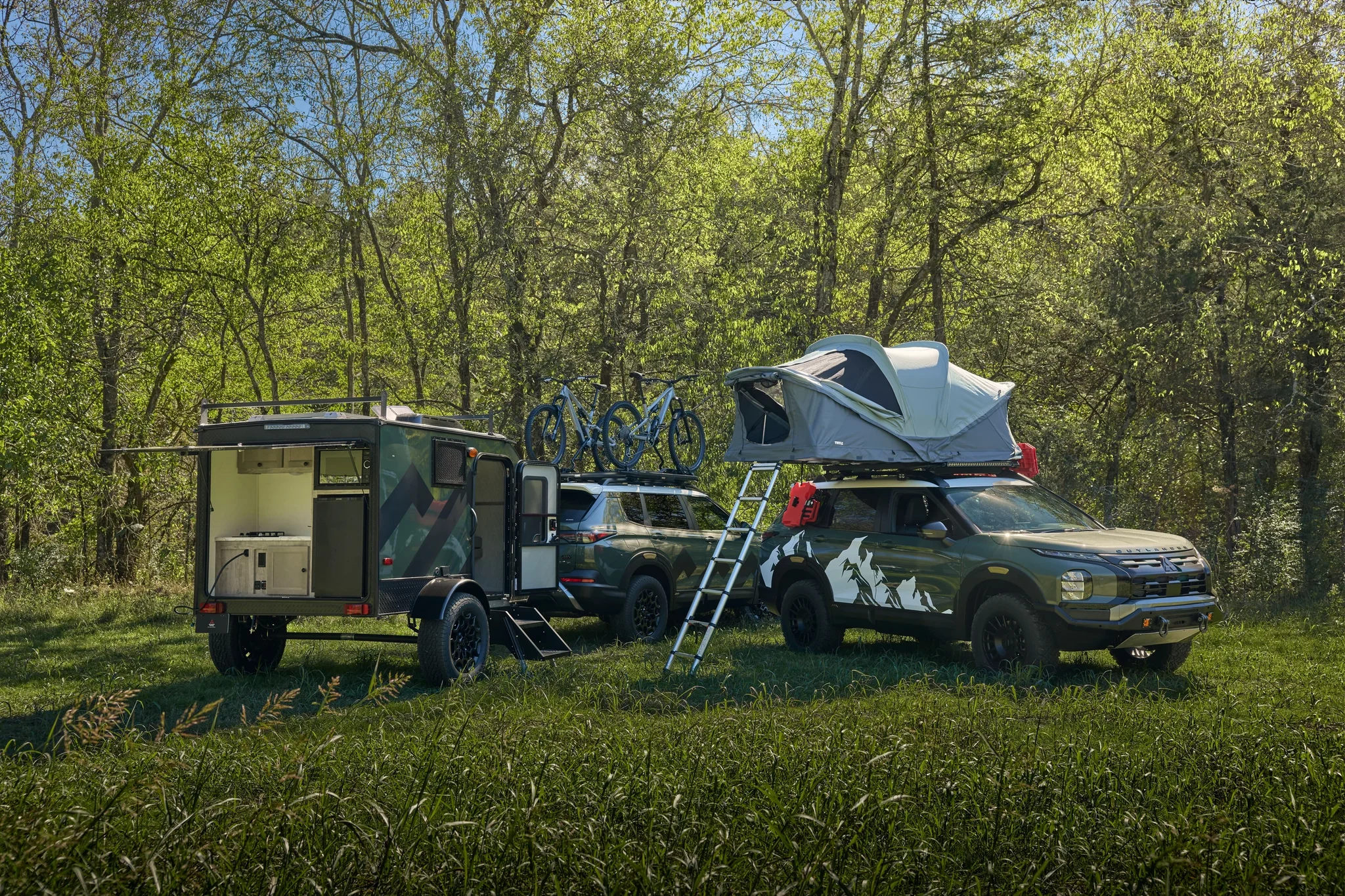 Dos Outlander Diamond Trail preparados con tienda de techo, remolque y bicicletas, en un campamento entre árboles