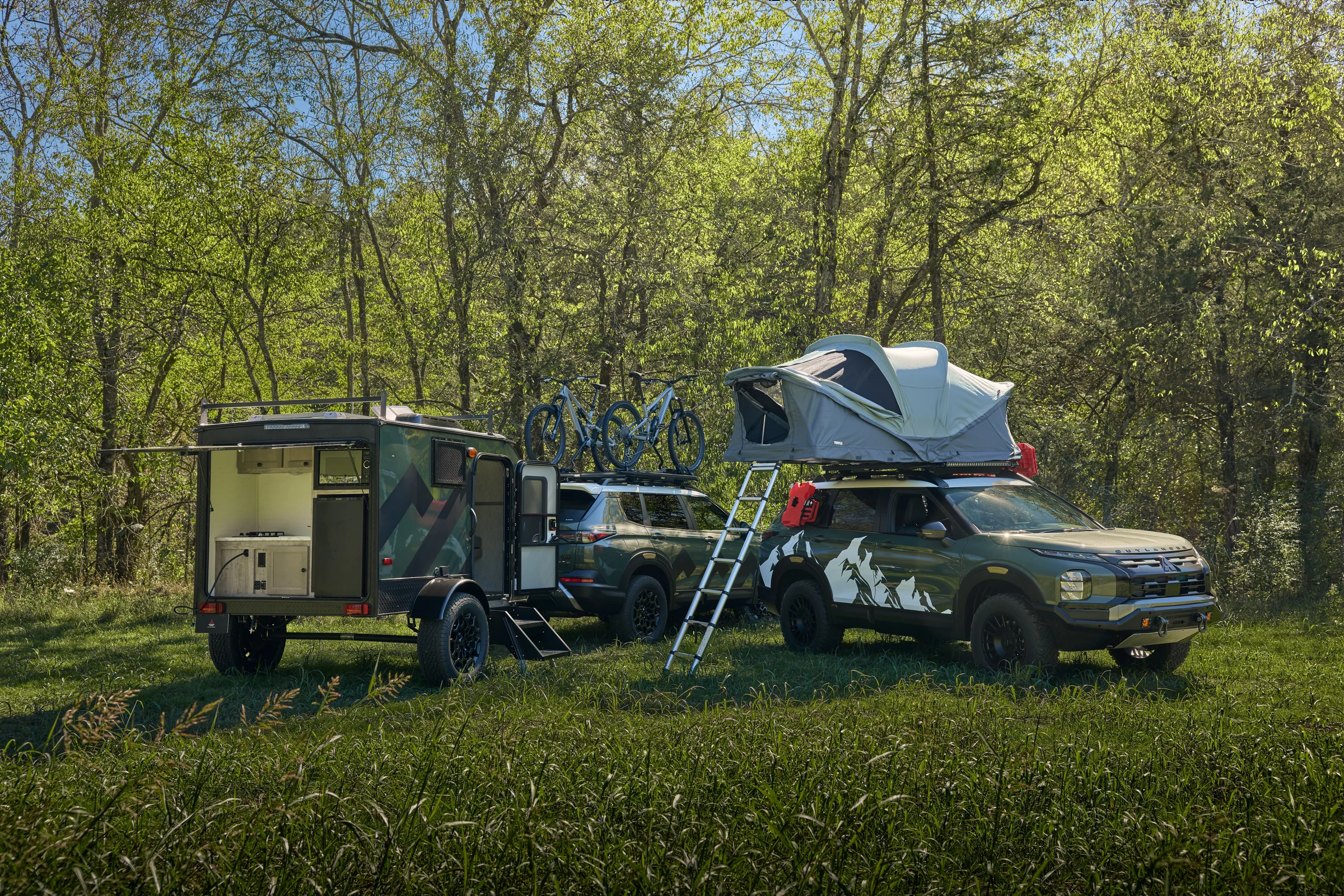 Dos Outlander Diamond Trail preparados con tienda de techo, remolque y bicicletas, en un campamento entre árboles