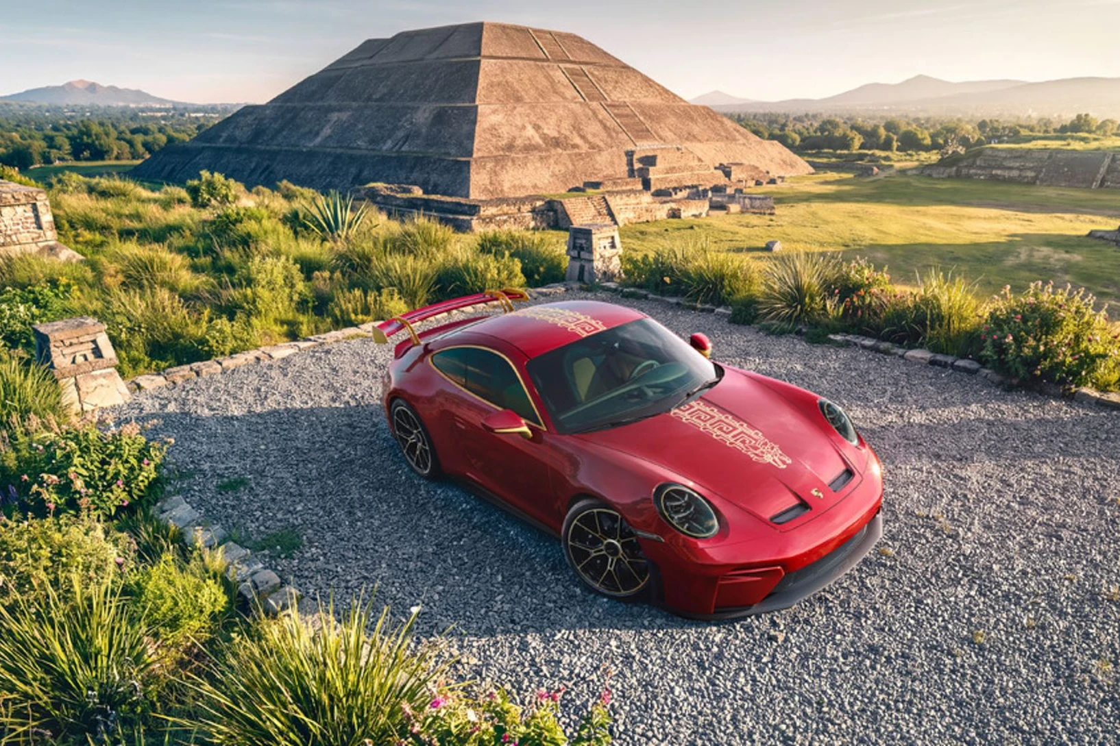 Porsche 911 GT3 «La Serpiente» rojo con motivos dorados, visto desde arriba en un camino de grava con una pirámide al fondo
