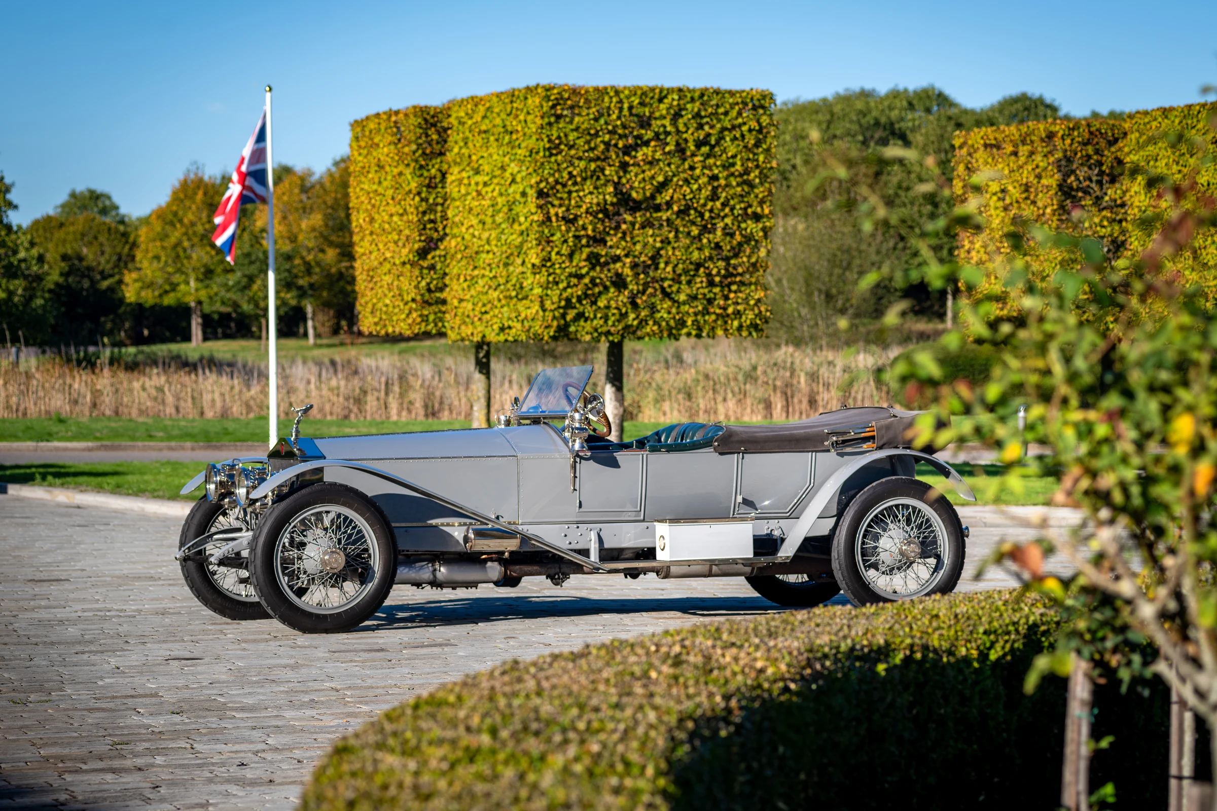 Rolls-Royce Silver Ghost con una bandera británica detrás, en un jardín con setos recortados y árboles