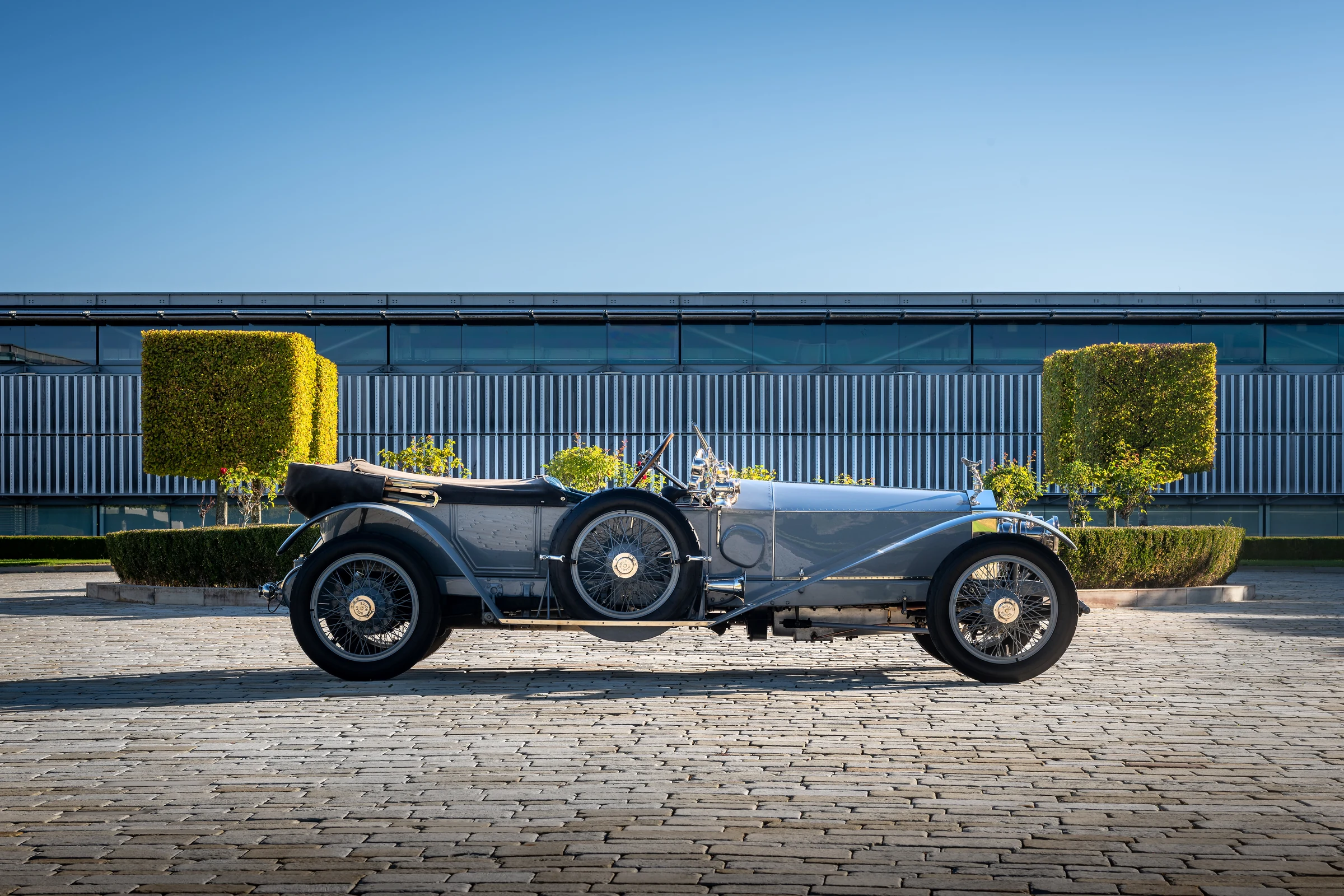 Perfil del Rolls-Royce Silver Ghost, un auto antiguo de carrocería abierta y ruedas de radios, ante un edificio de cristal