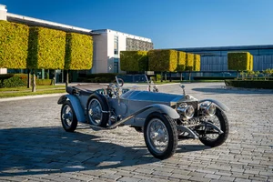 Rolls-Royce Silver Ghost plateado, descapotable con ruedas de radios, sobre adoquines frente a un edificio moderno