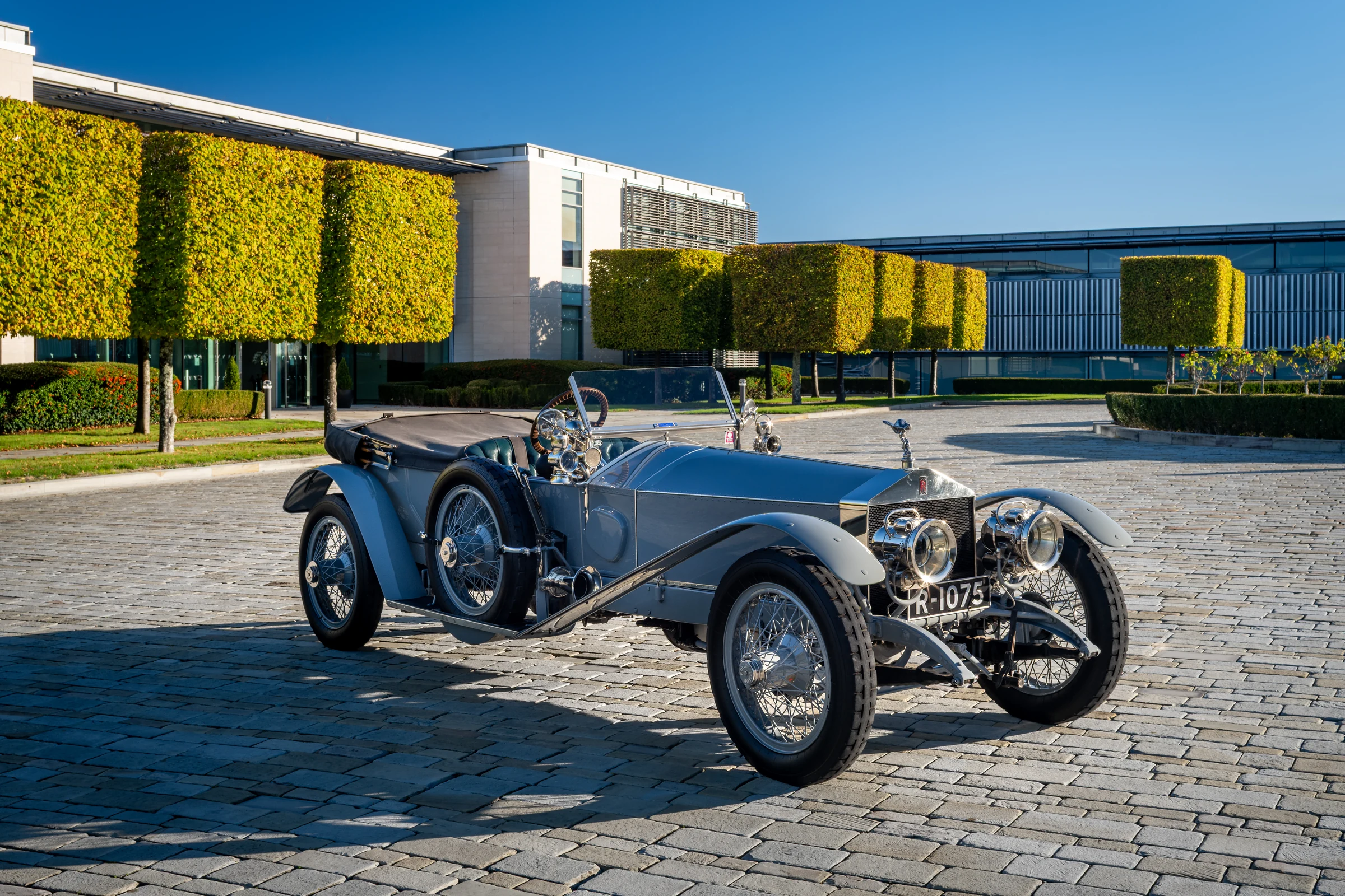 Rolls-Royce Silver Ghost plateado, descapotable con ruedas de radios, sobre adoquines frente a un edificio moderno