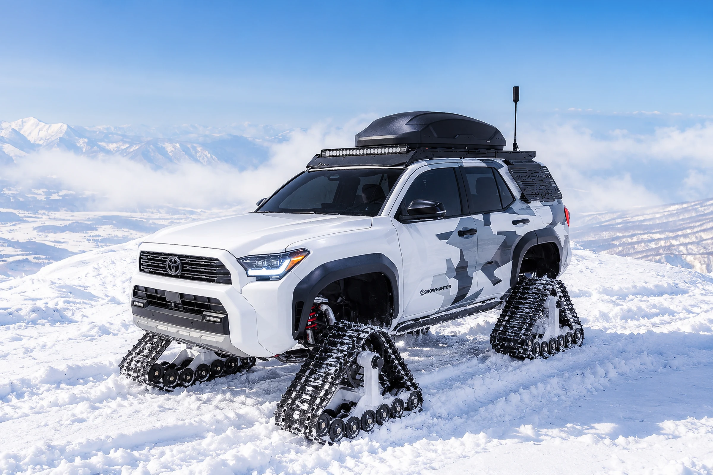 Toyota 4Runner Snowhunter blanca con camuflaje gris y orugas, de tres cuartos frontales, sobre nieve y con montañas atrás