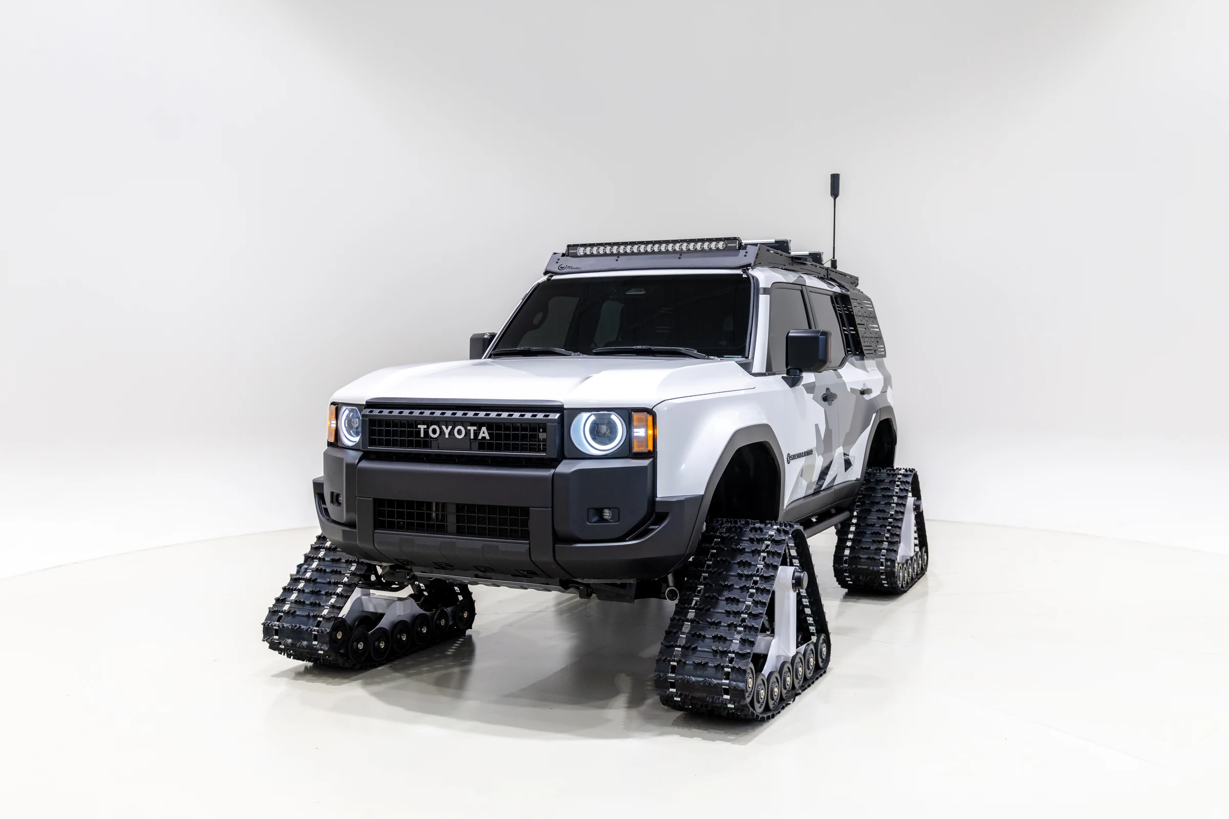Toyota Land Cruiser Snowhunter blanco con orugas negras y barra de luces, de tres cuartos frontales, en un estudio