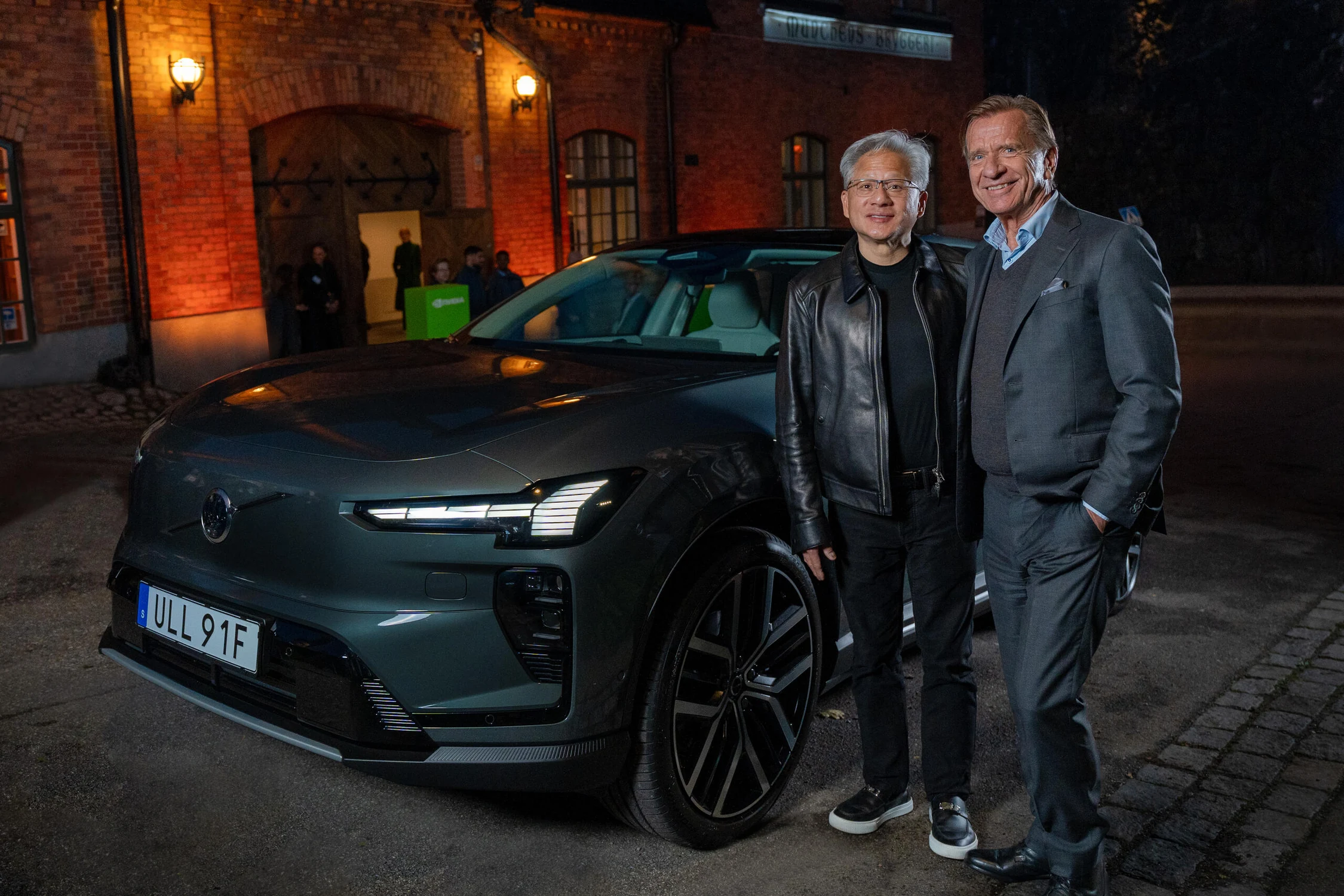 Jensen Huang, de NVIDIA, y Håkan Samuelsson, de Volvo Cars, junto a un Volvo eléctrico gris frente a un edificio de ladrillo
