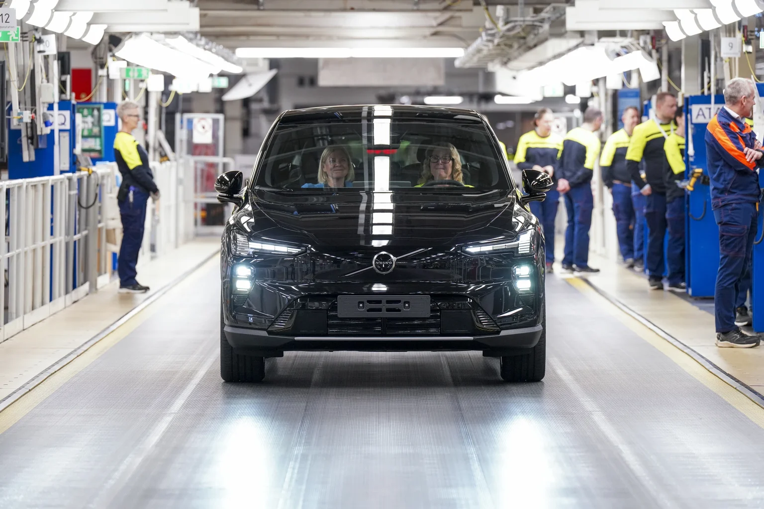 Un Volvo EX60 negro avanza por la línea de montaje de Volvo, con operarios de ropa amarilla y azul a los lados