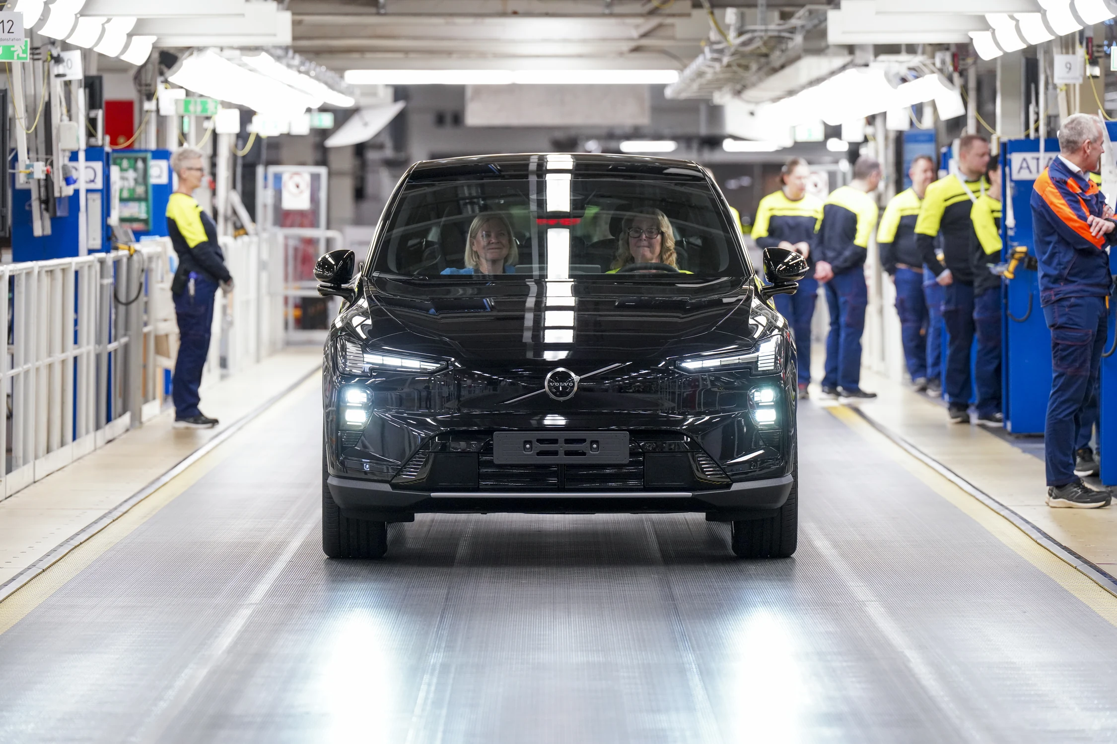 Un Volvo EX60 negro avanza por la línea de montaje de Volvo, con operarios de ropa amarilla y azul a los lados